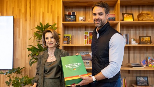 Raquel Teixeira e Eduardo Leite sorriem em p&eacute;, em uma sala com paredes e estantes de madeira. Juntos, seguram uma pasta verde com a inscri&ccedil;&atilde;o &ldquo;Educa&ccedil;&atilde;o &ndash; Rede Estadual&rdquo;.