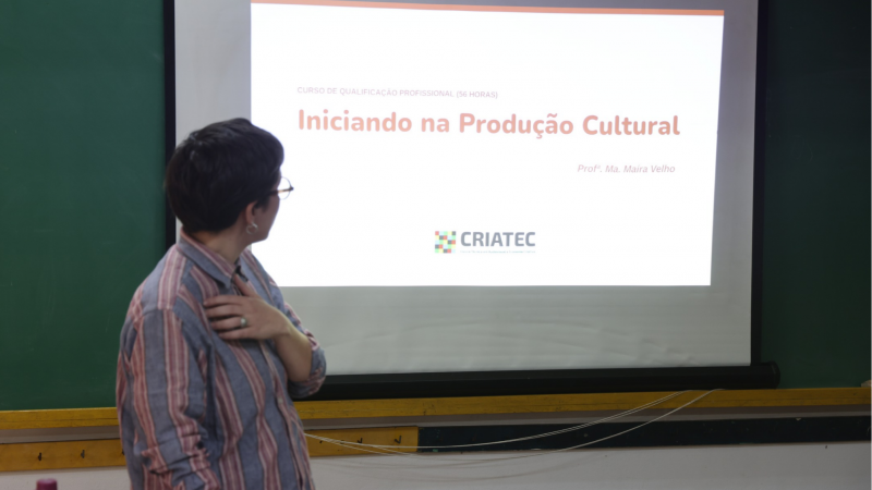 Professora Ma&iacute;ra aparece de costas olhando para uma tela de proje&ccedil;&atilde;o com um slide onde se l&ecirc; "Iniciando na Produ&ccedil;&atilde;o Cultural"