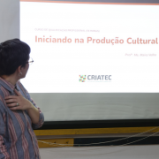 Professora Ma&iacute;ra aparece de costas olhando para uma tela de proje&ccedil;&atilde;o com um slide onde se l&ecirc; "Iniciando na Produ&ccedil;&atilde;o Cultural"
