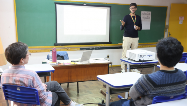 Superintendente da Educação Profissional, Tom Collier, aparece em pé gesticulando na sala de aula, enquanto a professora e um estudante aparecem sentados nas cadeiras olhando para ele.