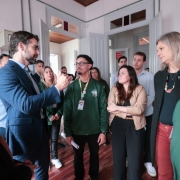 Foto de um grupo de pessoas no interior do col&eacute;gio. &Agrave; esquerda, o governador Eduardo Leite veste um terno azul e gesticula enquanto fala para os demais, que prestam aten&ccedil;&atilde;o. Entre os ouvintes, nota-se pessoas vestindo jaquetas verdes de uniforme da Rede Estadual. O local possui portas brancas e piso de madeira.