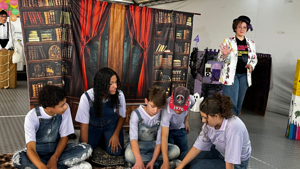 Cinco estudantes aparecem sentados em um tapete no ch&atilde;o interagindo com um livro. Uma menina de chap&eacute;u est&aacute; em p&eacute; ao lado deles. Ao fundo, um cen&aacute;rio de pano com imagem de uma estante cheia de livros e uma janela.